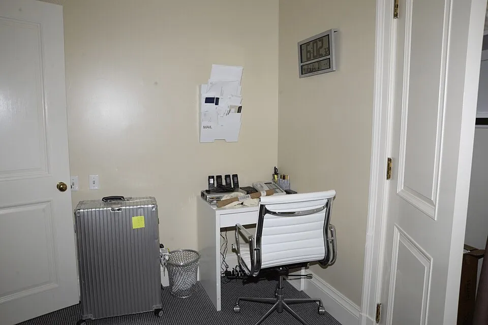 EFTA00000817 - Small workspace with a white desk office chair silver suitcase mail holder and a digital clock on the wall.jpg